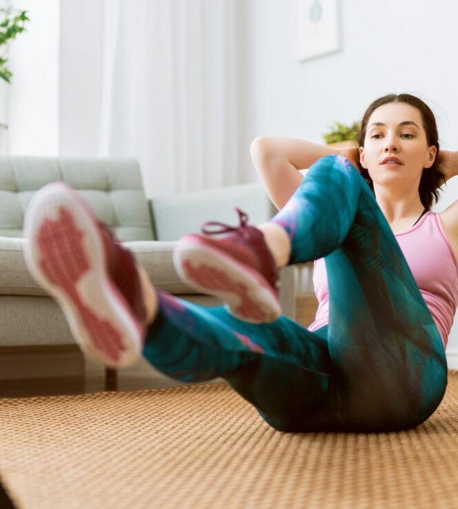 Giovane donna in abbigliamento sportivo si allena a corpo libero nel salotto di casa seguendo una piattaforma di fitness online con il proprio iPad.